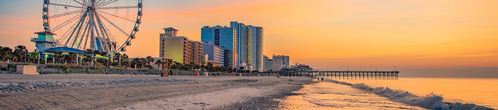 A seaside sunset with a large ferris wheel, high-rise hotels, and a long pier along the shore.