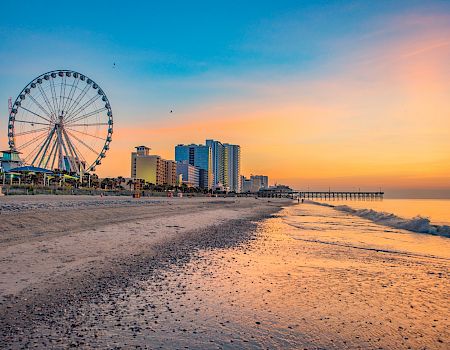 A seaside sunset with a large ferris wheel, high-rise hotels, and a long pier along the shore.