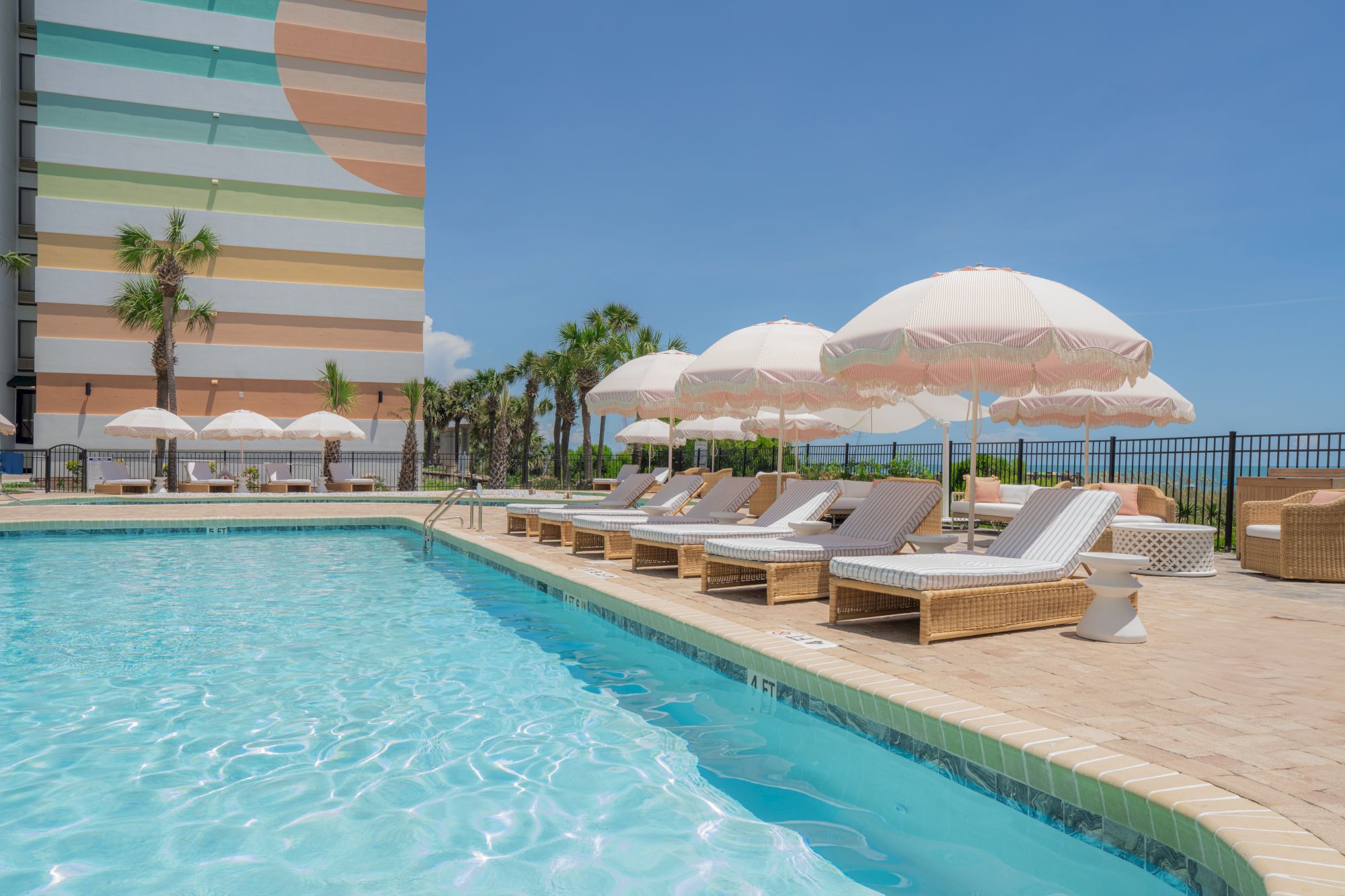 A vibrant poolside scene with wicker loungers, pink umbrellas, and a colorful building, set against a clear blue sky.