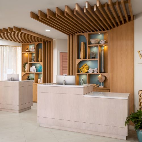 The image shows a modern hotel reception area with wooden decor, two counters, decorative items on shelves, and the "VOCO by IHG" logo on the wall.
