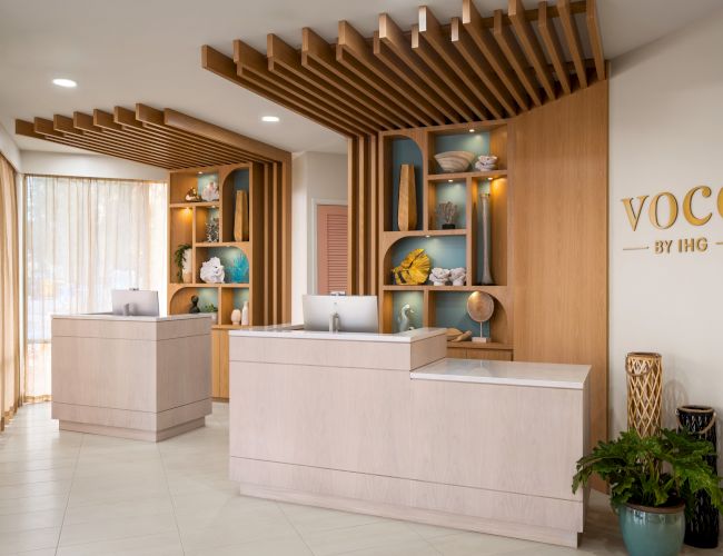 The image shows a modern hotel reception area with wooden decor, two counters, decorative items on shelves, and the "VOCO by IHG" logo on the wall.