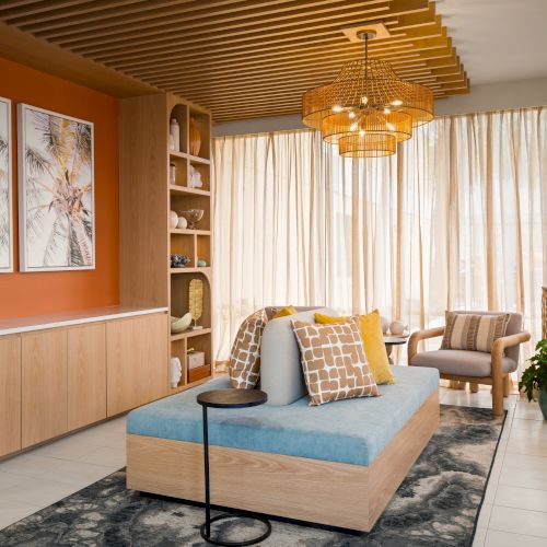 A modern living room with a wooden ceiling, artwork, comfy seating, warm lighting, plants, and sheer curtains enhancing the natural light.