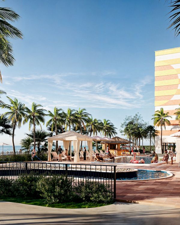 A tropical resort scene with palm trees, a pool, people relaxing, and a tall building, set against a clear, sunny sky.