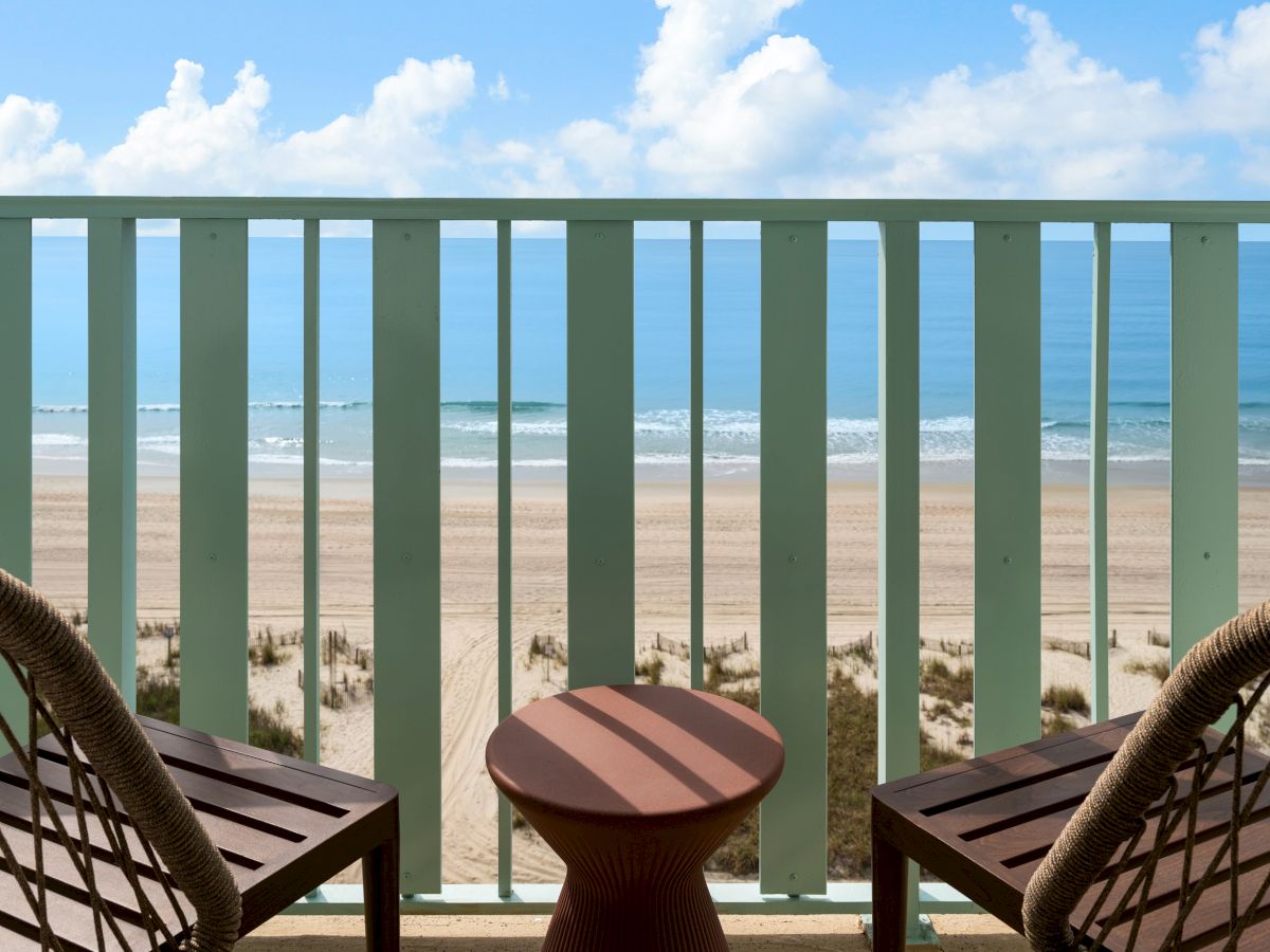 A beach view with two chairs and a small table on a balcony overlooking the ocean under a blue sky with clouds.