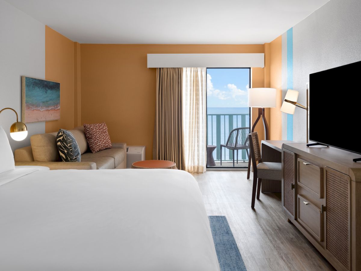 A modern hotel room with a bed, sofa, TV, and balcony view. Orange and blue accents decorate the space, creating a cozy atmosphere.