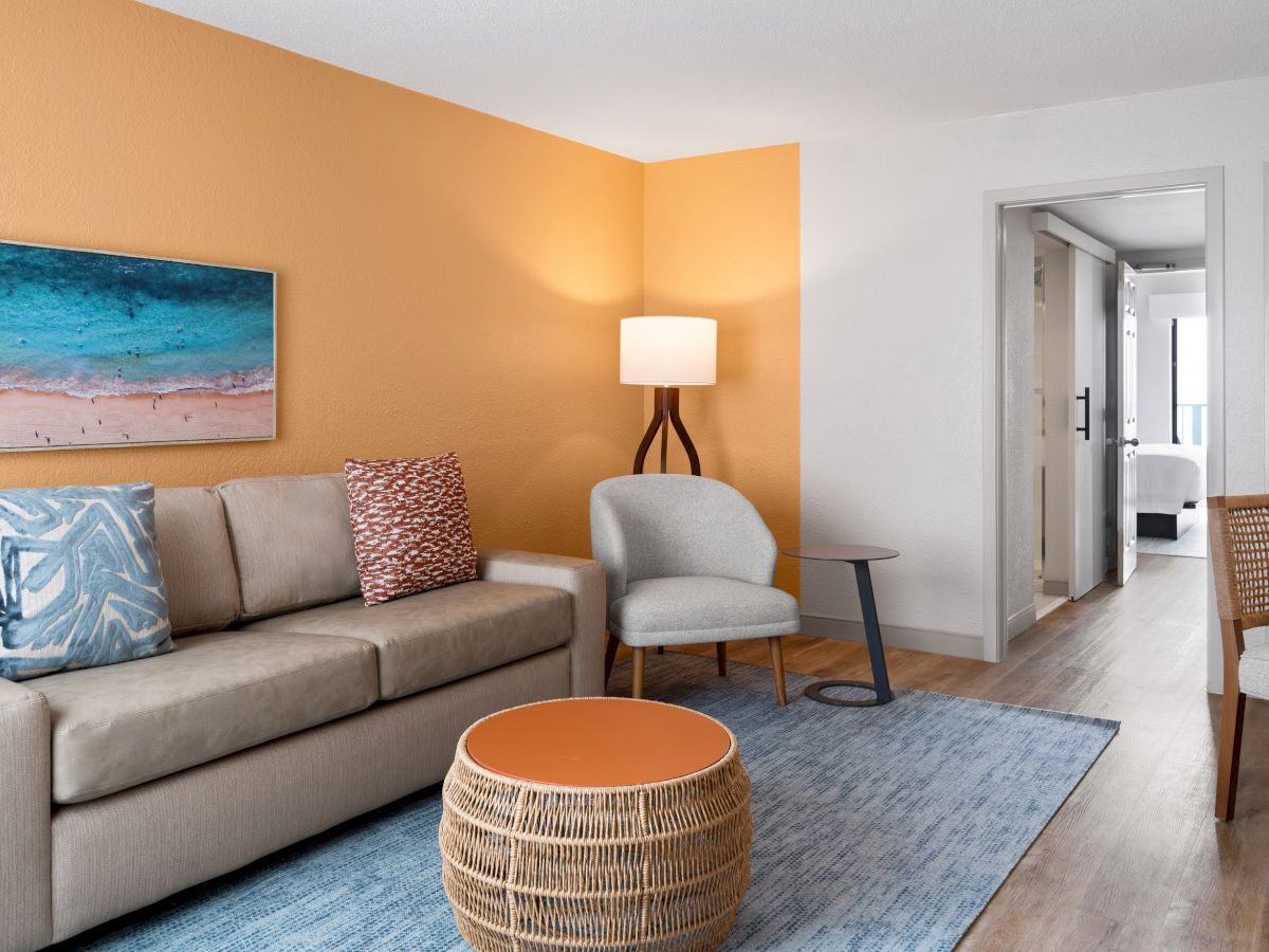 A cozy living room with a beige sofa, colorful cushions, a wicker coffee table, and a lamp. The walls are orange and white with a beach painting.