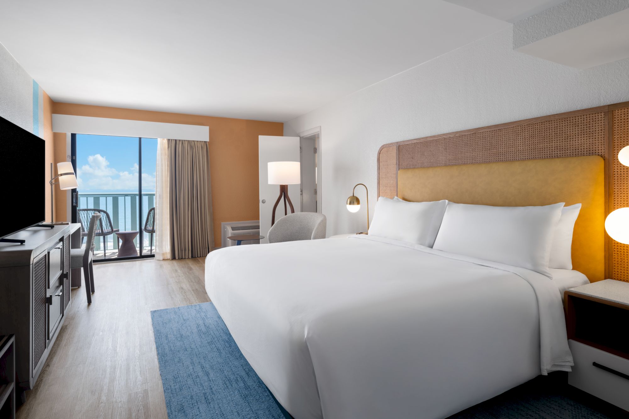 A hotel room with a large bed, TV, lamps, and balcony view of the ocean under a blue sky.