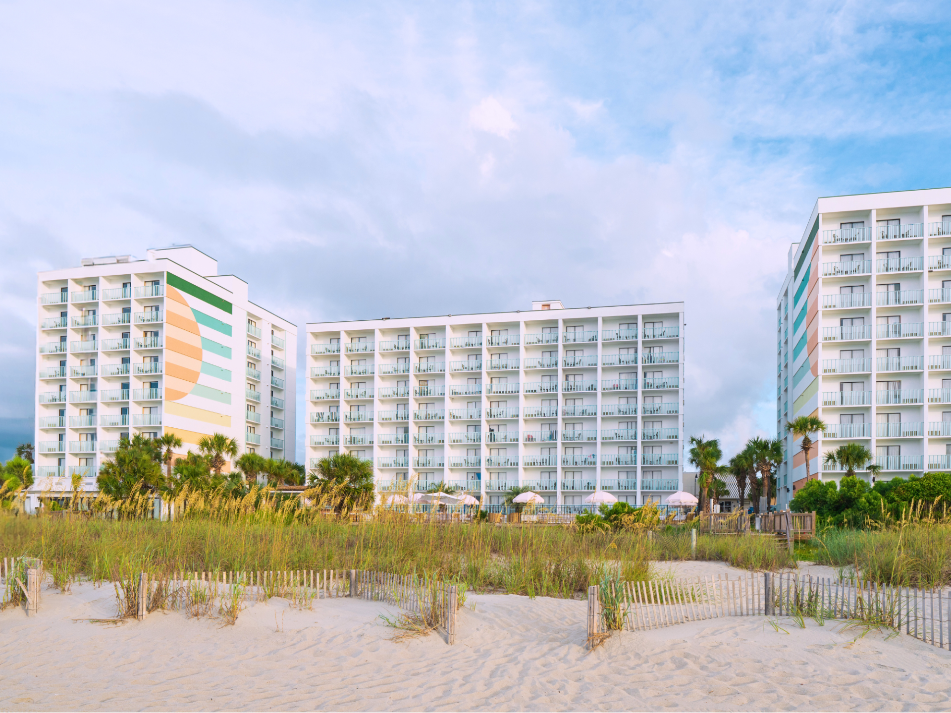 The image shows three beachfront hotels with colorful patterns, set against sandy dunes and a partly cloudy sky.