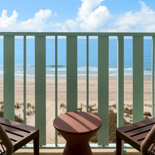 Two chairs and a small table on a balcony overlook a sandy beach and the ocean under a blue sky with scattered clouds.