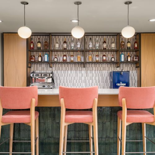 A bar area with pink chairs, pendant lights, and a counter with bottles, a coffee machine, and a blue bag displayed on the shelves.