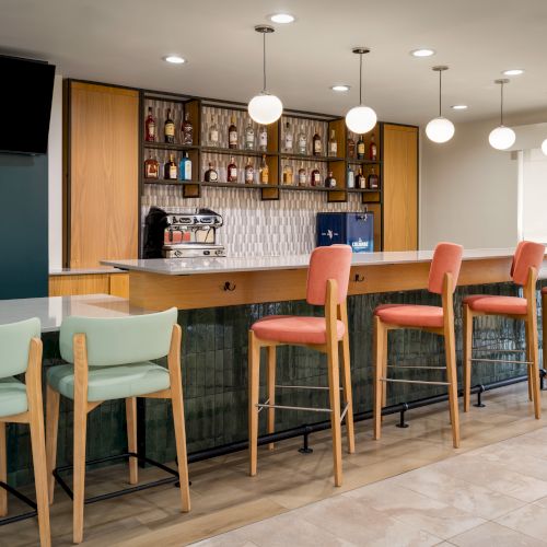 The image shows a modern bar with colorful chairs, a countertop, shelving with bottles, and hanging globular lights in a sleek setting.