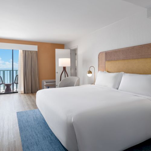 A modern hotel room with a large bed, TV, and balcony overlooking the ocean. Bright and inviting space with stylish decor.