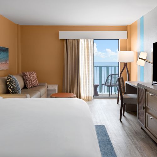 A modern hotel room with a bed, sofa, TV, and balcony overlooking the ocean, featuring orange and neutral decor tones.