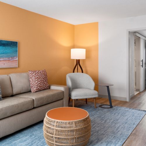 A cozy living room with a beige sofa, orange accent wall, beach artwork, and a wicker coffee table, leading into a bedroom.
