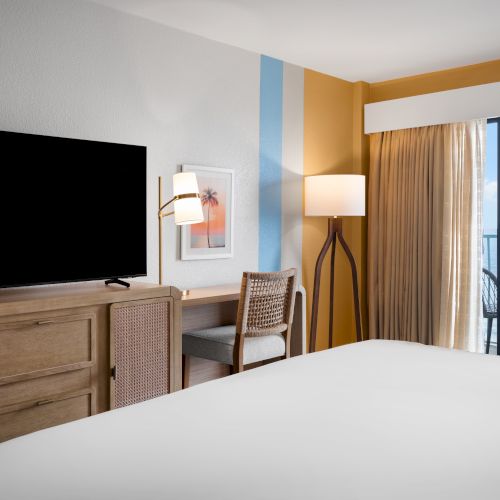 The image shows a hotel room with a bed, a flat-screen TV on a wooden stand, a chair, a floor lamp, and a balcony with a view of the ocean.