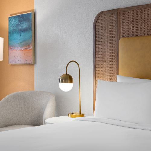 A hotel room features a bed with white linens, a modern lamp, a comfortable chair, and a colorful painting on the wall.
