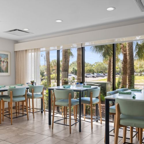 Modern dining room with green chairs and set tables, big windows showing palm trees and parked cars outside.