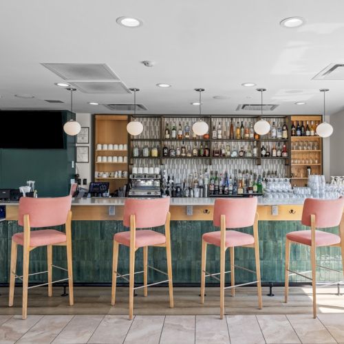A modern hotel bar with pink stools, a stocked liquor shelf, glassware, and a green tile counter.