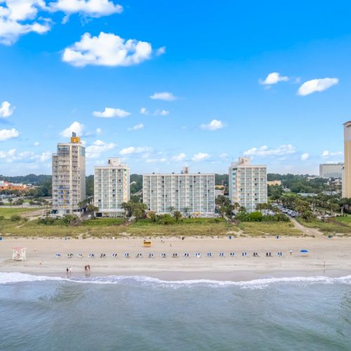 A sandy beach with rows of umbrellas and shoreline waves, backed by tall apartment/hotel buildings under a blue sky.