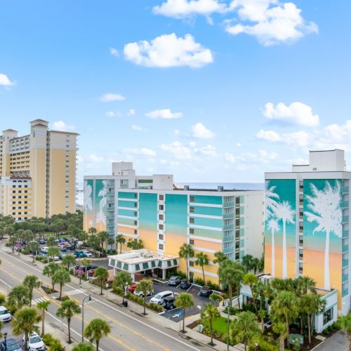 A sunny view of a beachfront hotel complex with colorful buildings, palm trees, and a busy parking lot under blue skies.