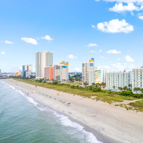 A sunny beachfront with a long sandy shore, ocean waves, and colorful beachfront hotels under a blue sky with clouds.