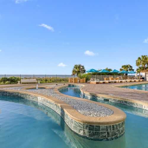 A sunny hotel pool with lounge chairs, blue umbrellas, and palm trees by the ocean under a clear sky.
