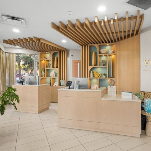 A modern hotel reception with &ldquo;voco by IHG&rdquo; branding, wood slats, and a central front desk with decor and plants.