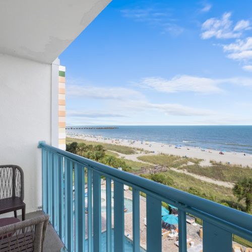 Balcony with a chair overlooks a sandy beach, ocean waves, and a bright blue sky.