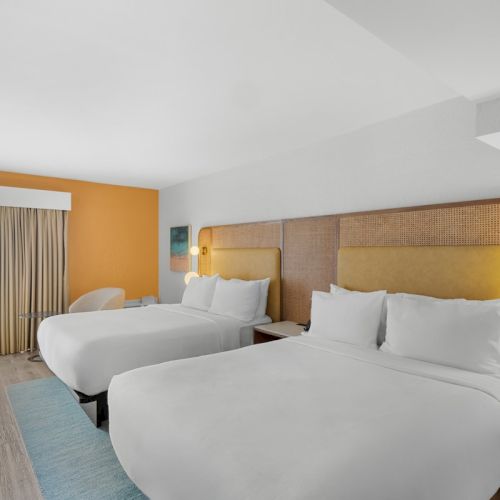 Bright hotel room with two double beds, white linens, and rattan headboards near a balcony door.