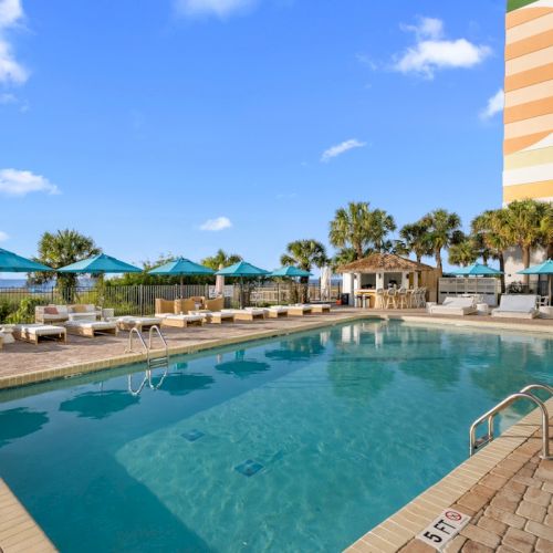 A resort outdoor pool with blue water, loungers, colorful umbrellas, palm trees, and the ocean under a sunny sky.