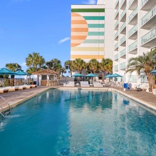 An outdoor hotel pool with lounge chairs and teal umbrellas beside modern buildings under a bright blue sky.