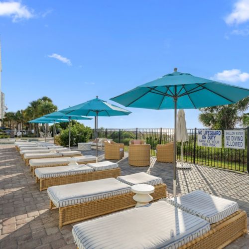 Resort poolside with neatly lined lounge chairs and green umbrellas under a bright blue sky, beside a black fence.