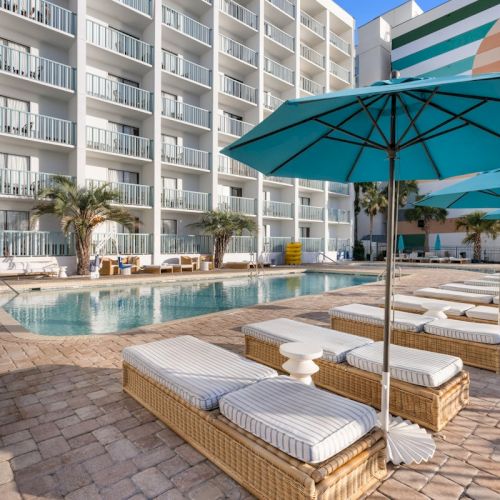 A resort hotel courtyard with a swimming pool, lounge chairs under green umbrellas, and palm trees beside a multi-story building.