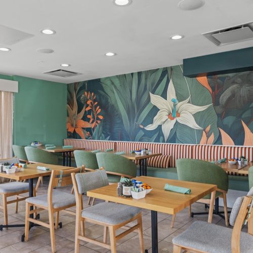 A bright caf&eacute; dining area with wooden tables, green upholstered chairs, and a tropical mural wall with flowers.