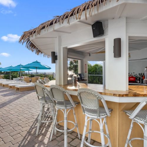 An outdoor seaside bar with wicker chairs, blue umbrellas, and a service counter under a thatched roof.