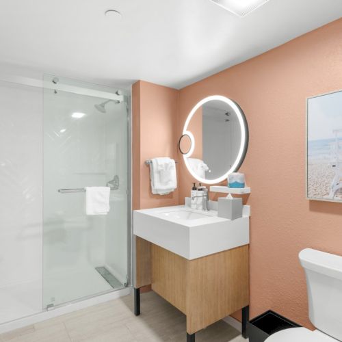 A modern bathroom with a glass shower, white sink vanity, round lit mirror, beach wall art, and a toilet.
