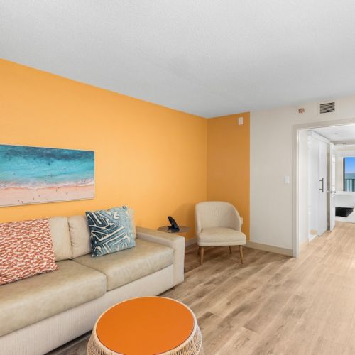 Bright living room with beige sofa, orange table, beach painting, and open door to hallway.