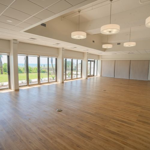 A spacious, empty room with wooden flooring, large windows, and modern lighting, offering views of greenery outside.
