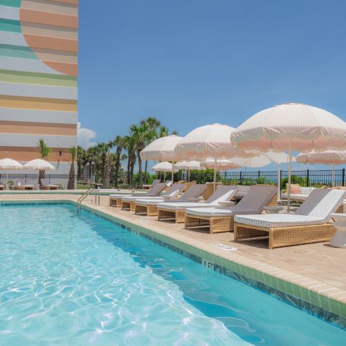 A poolside scene with wicker loungers, white and pink umbrellas, palm trees, and a colorful building under a clear blue sky.