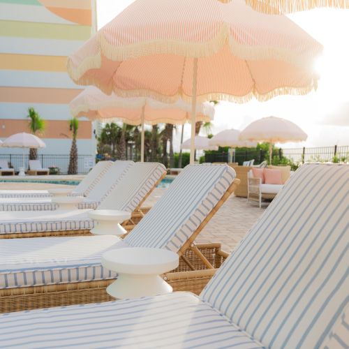 Poolside view with striped lounge chairs and pink umbrellas. A pastel-colored building and palm trees are in the background.