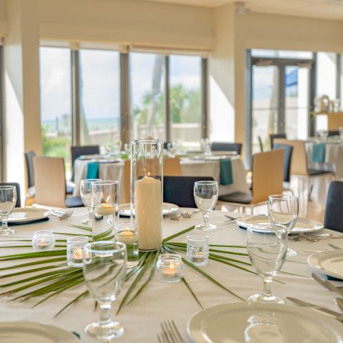 An elegantly set dining table with glassware, plates, candles, and palm leaves in a bright room with large windows and a view.