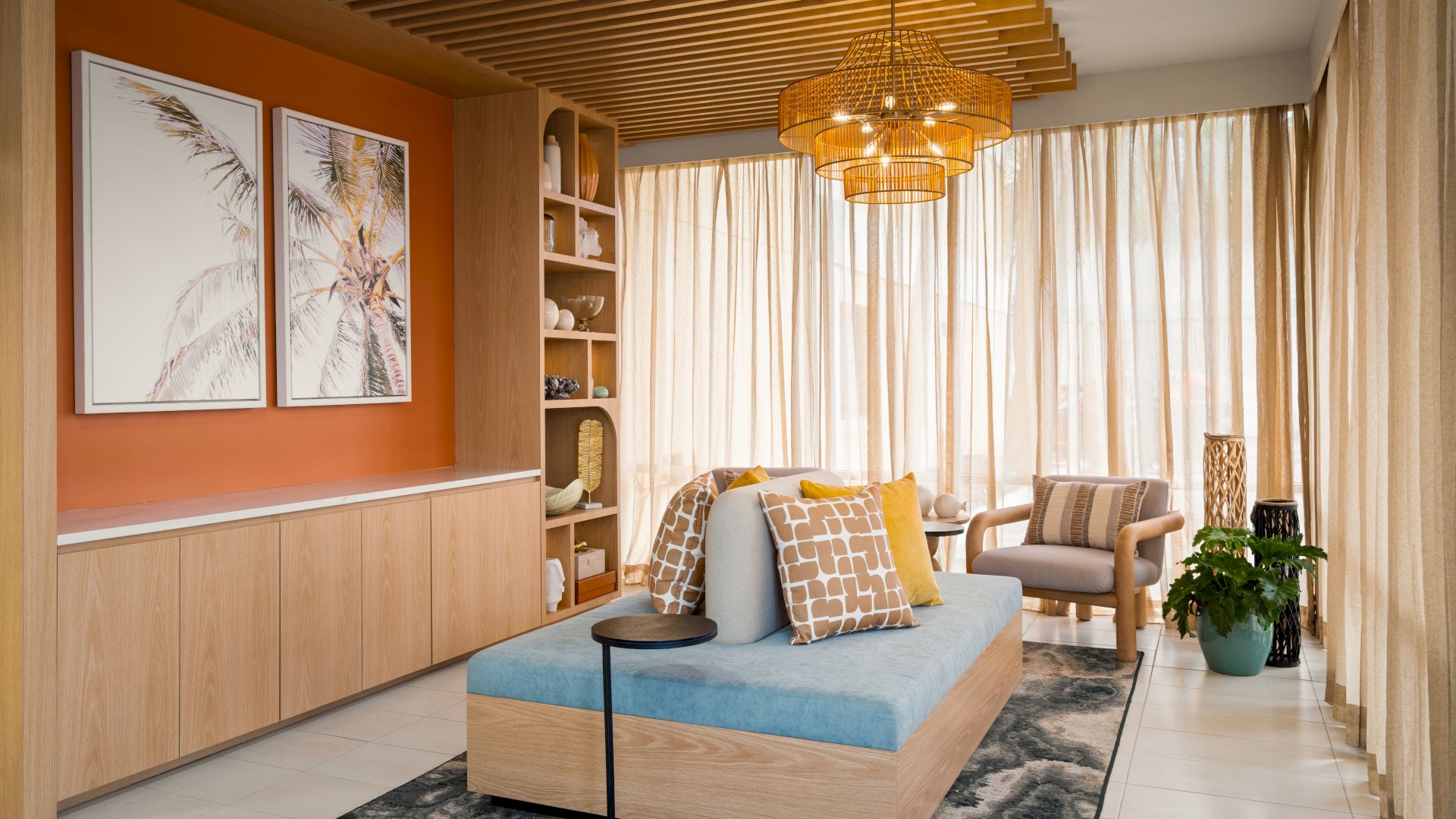 A cozy living room with wood accents, large windows, a sofa, armchair, art pieces, potted plants, and a patterned rug on the floor.