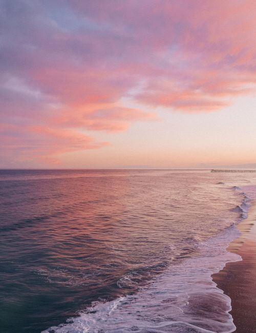 A serene beach scene at sunset with pink clouds, gentle waves, and a distant city skyline along the coast.