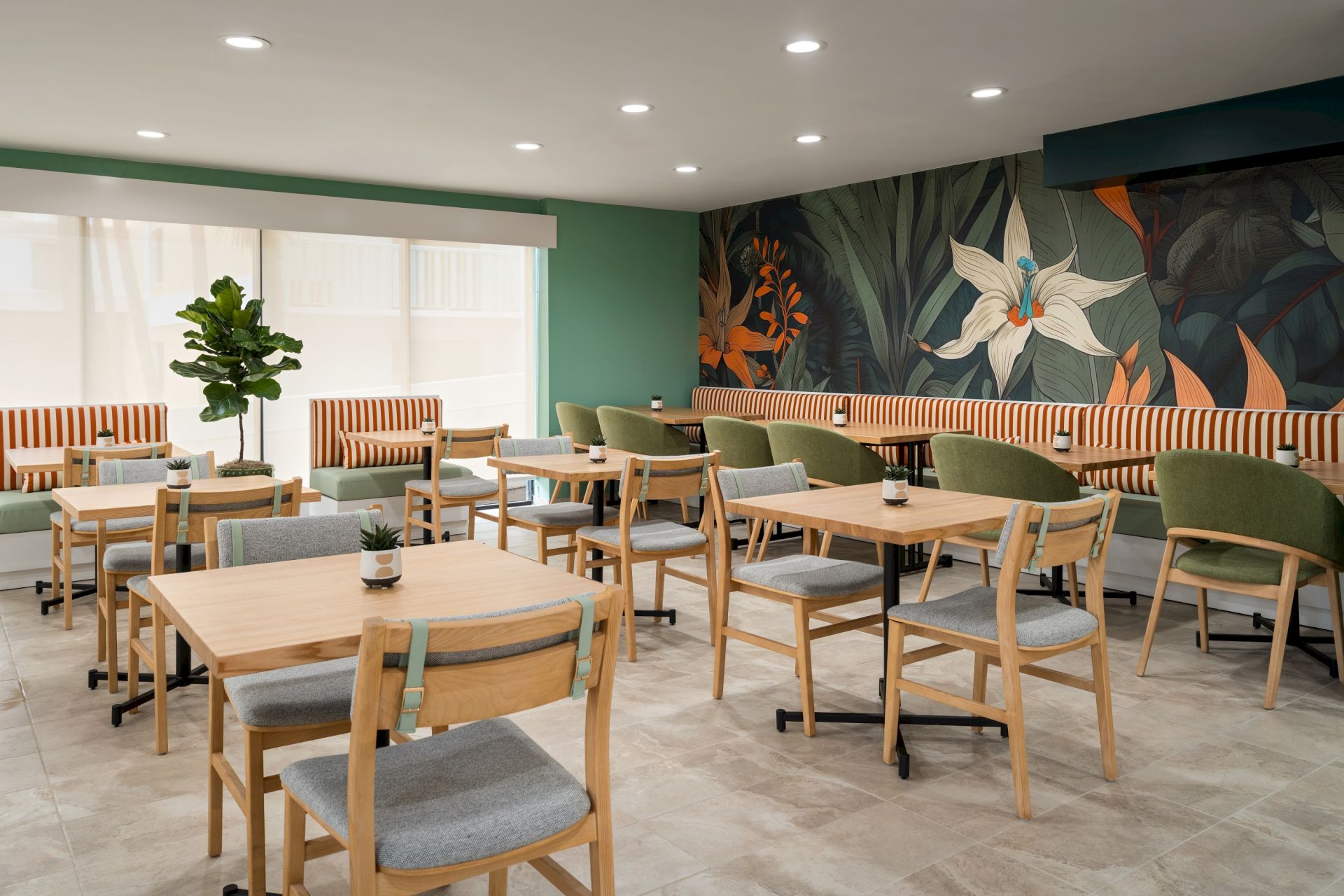 A modern caf&eacute; with wooden tables, green chairs, wall art with floral design, and bright lighting. There&rsquo;s a small potted plant on a table.