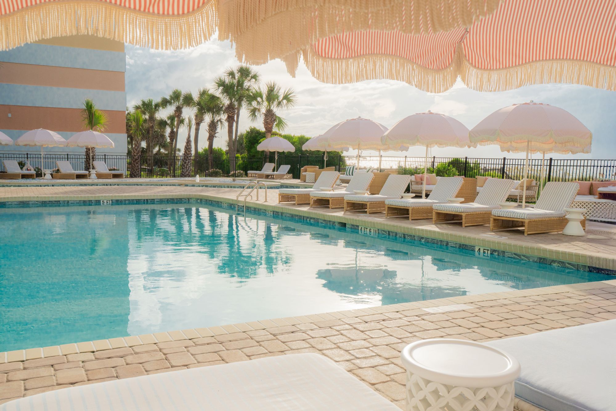 A serene poolside scene with lounge chairs, pink umbrellas, palm trees, and a building in the background under a striped awning.