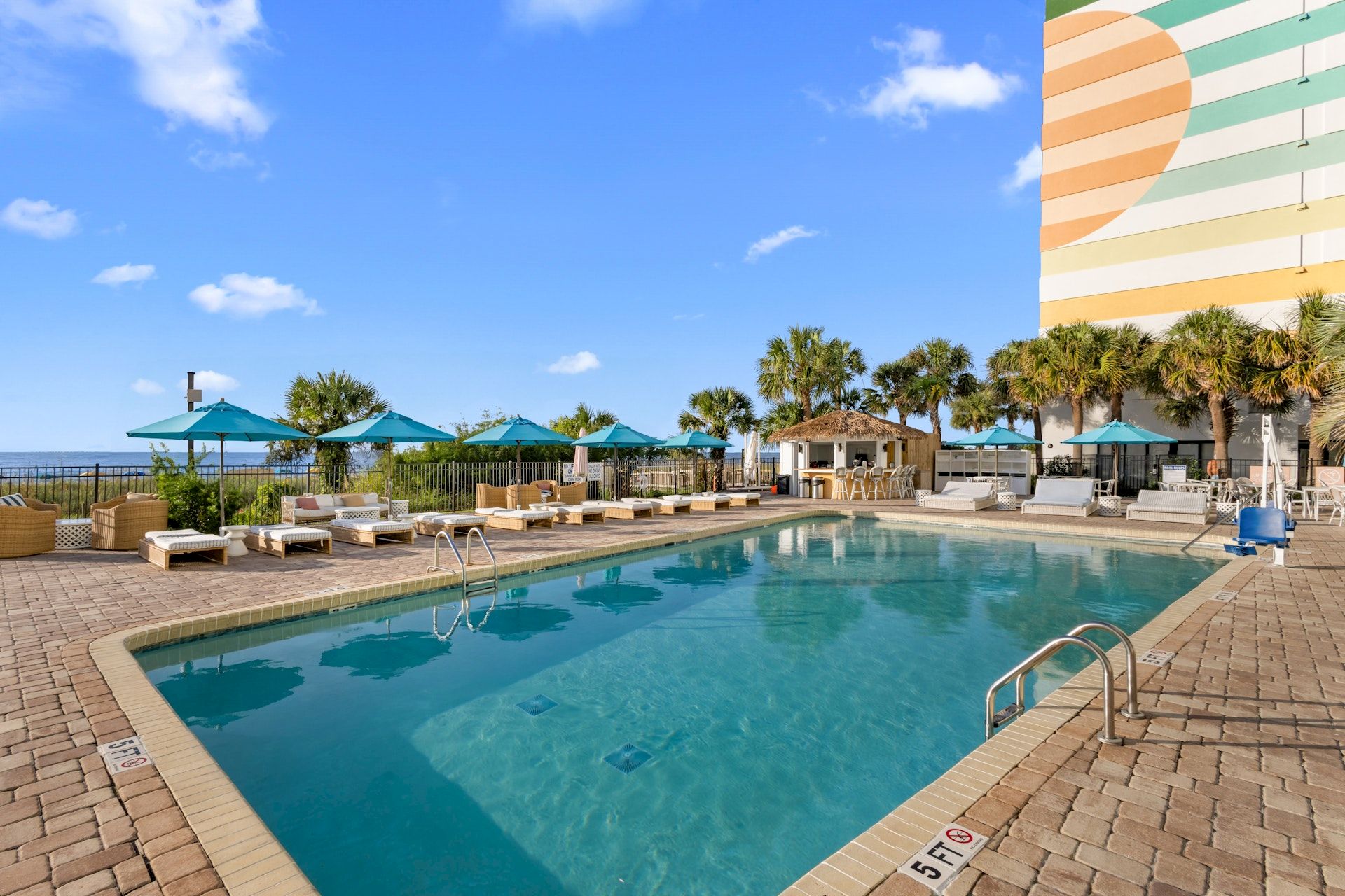 A sunny hotel pool with lounge chairs and teal umbrellas beside the ocean under a bright blue sky.