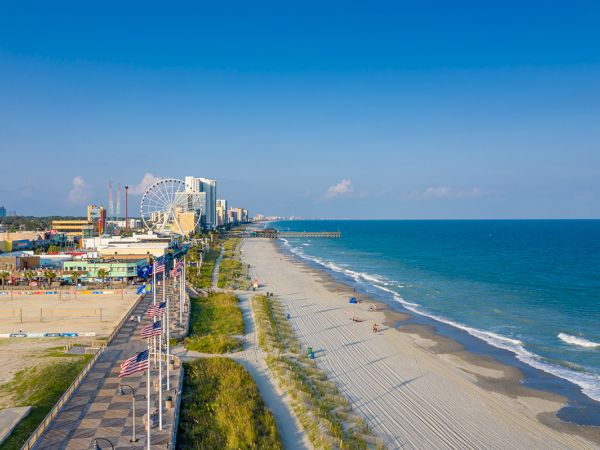 A beach scene with a boardwalk, flags, hotels, a ferris wheel, and a sandy shoreline meeting the ocean under a clear blue sky.