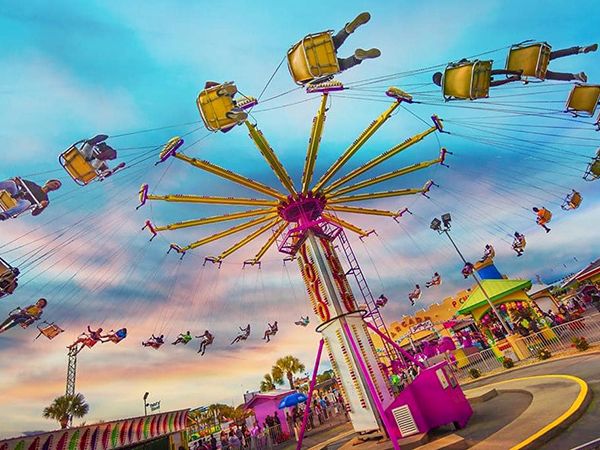 The image shows a colorful amusement park with people enjoying a spinning swing ride under a vibrant sky.
