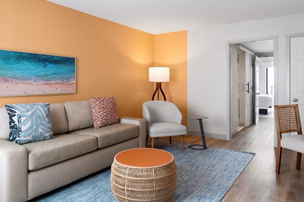 A cozy living room with a beige sofa, colorful pillows, an orange accent wall, a wicker coffee table, and a floor lamp. Blue rug included.
