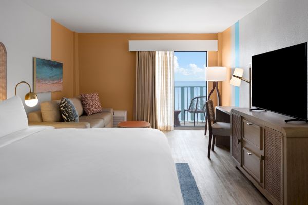 A modern hotel room with a bed, sofa, TV, and balcony door opening to a view. The decor includes warm tones and a floor lamp.